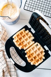 A waffle iron with two cooked waffles in it. Behind the iron is a blunder filled with waffle batter next to it as part of a black wire rack.