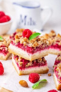 Two raspberry cheesecake bars stacked on top of one another on a piece of white parchment paper. There are parts of raspberry cheesecake bars surrounding them.