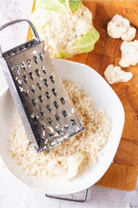 Cutting board with a head of cauliflower, three cauliflower florettes, and a white bowl filled with riced cauliflower. There is a box grater inside the white bowl.