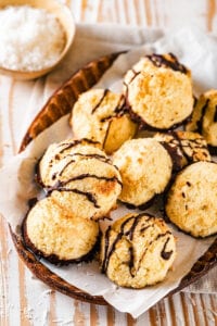 An oval brown bowl lined with white parchment paper with a bunch of coconut macaroons on top. Some of the macaroons have chocolate on the bottom of them and some have chocolate drizzled on top. Behind the bowl is a small bowl of shredded coconut.
