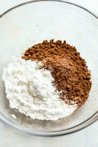 Cocoa powder, sweetener, and protein powder, in a glass bowl. The ingredients are unmixed.