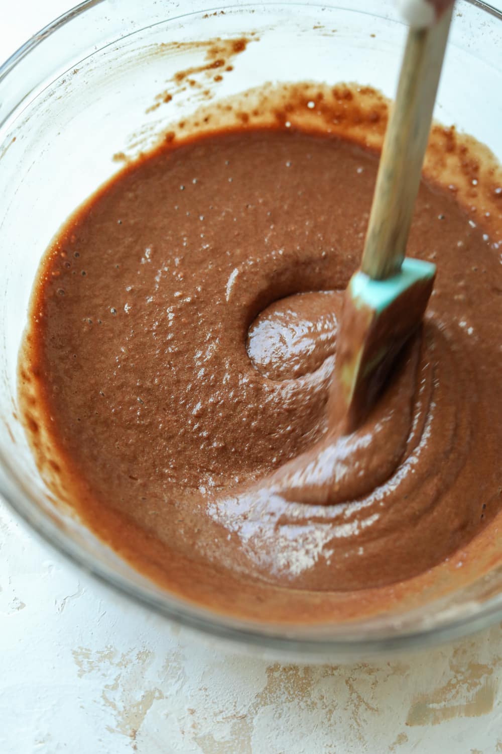 A spatula mixing chocolate cake batter in a glass bowl.