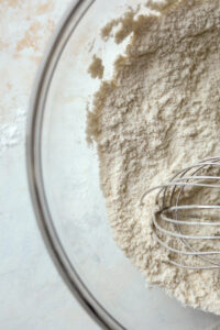 A clear bowl filled with flour and a wire whisk.