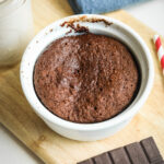 Chocolate cake in a white mug that is set on a wooden cutting board surrounded by a glass of milk, a blue napkin, a red and white straw, and a piece of chocolate.