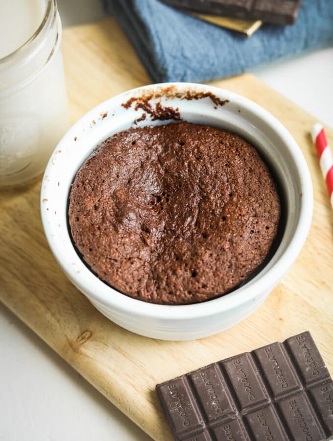 Chocolate cake in a white mug that is set on a wooden cutting board surrounded by a glass of milk, a blue napkin, a red and white straw, and a piece of chocolate.