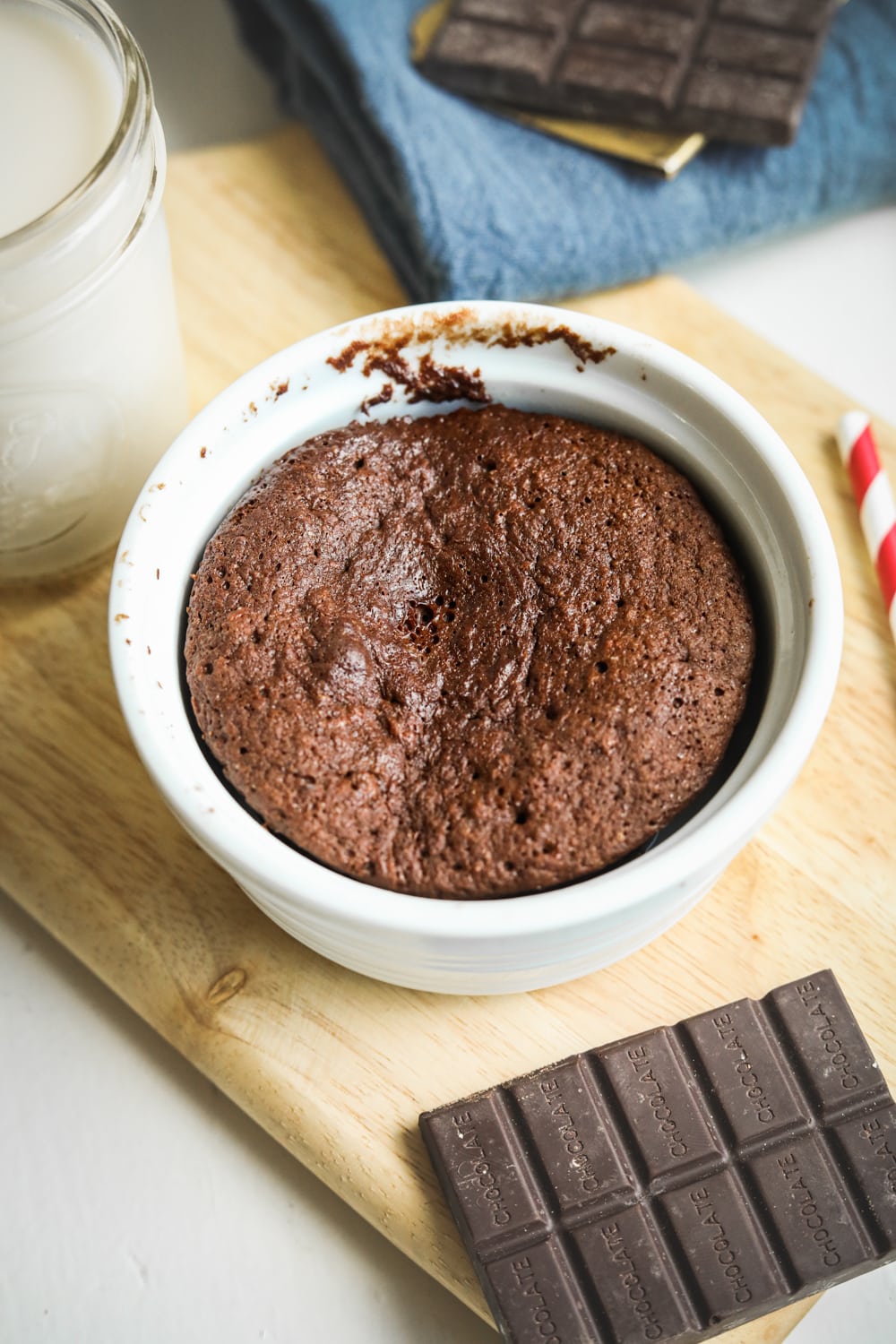 Chocolate cake in a white mug that is set on a wooden cutting board surrounded by a glass of milk, a blue napkin, a red and white straw, and a piece of chocolate.