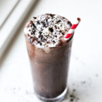 A glass full a chocolate shake. The shake is topped with whipped cream and black cocoa powder.