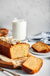 Part of a loaf of banana bread and a piece of white parchment paper on a wooden cutting board. A slice of banana bread is cut out and it is lying face down in front of the cutting Board. Behind the low is a glass of milk and a white plate with a slice of banana bread on it.