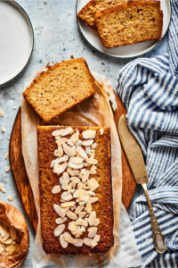 A loaf of banana bread and a piece of parchment paper on a wooden cutting board. There's a slice lying facedown in front of the loaf and in front of that is part of a white plate with two slices of banana bread on top of one another.