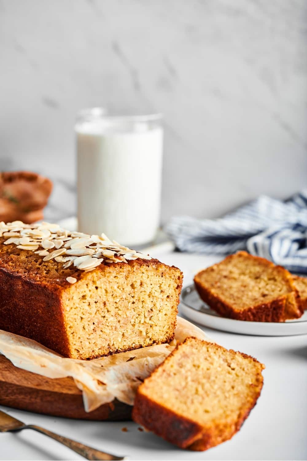 Part of a loaf of banana bread and a piece of white parchment paper on a wooden cutting board. A slice of banana bread is cut out and it is lying face down in front of the cutting Board. Behind the low is a glass of milk and a white plate with a slice of banana bread on it.