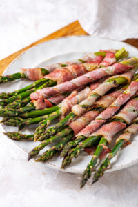 White plate on boarding cutting board on top of a white counter with bacon wrapped asparagus on the plate.