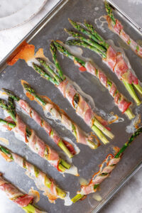 A few pieces of asparagus wrapped in bacon and a few bundles of asparagus wrapped in bacon on a baking sheet.