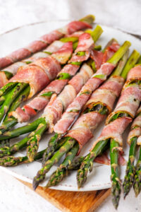 A white plate on a wooden cutting board with bacon wrapped asparagus on it.