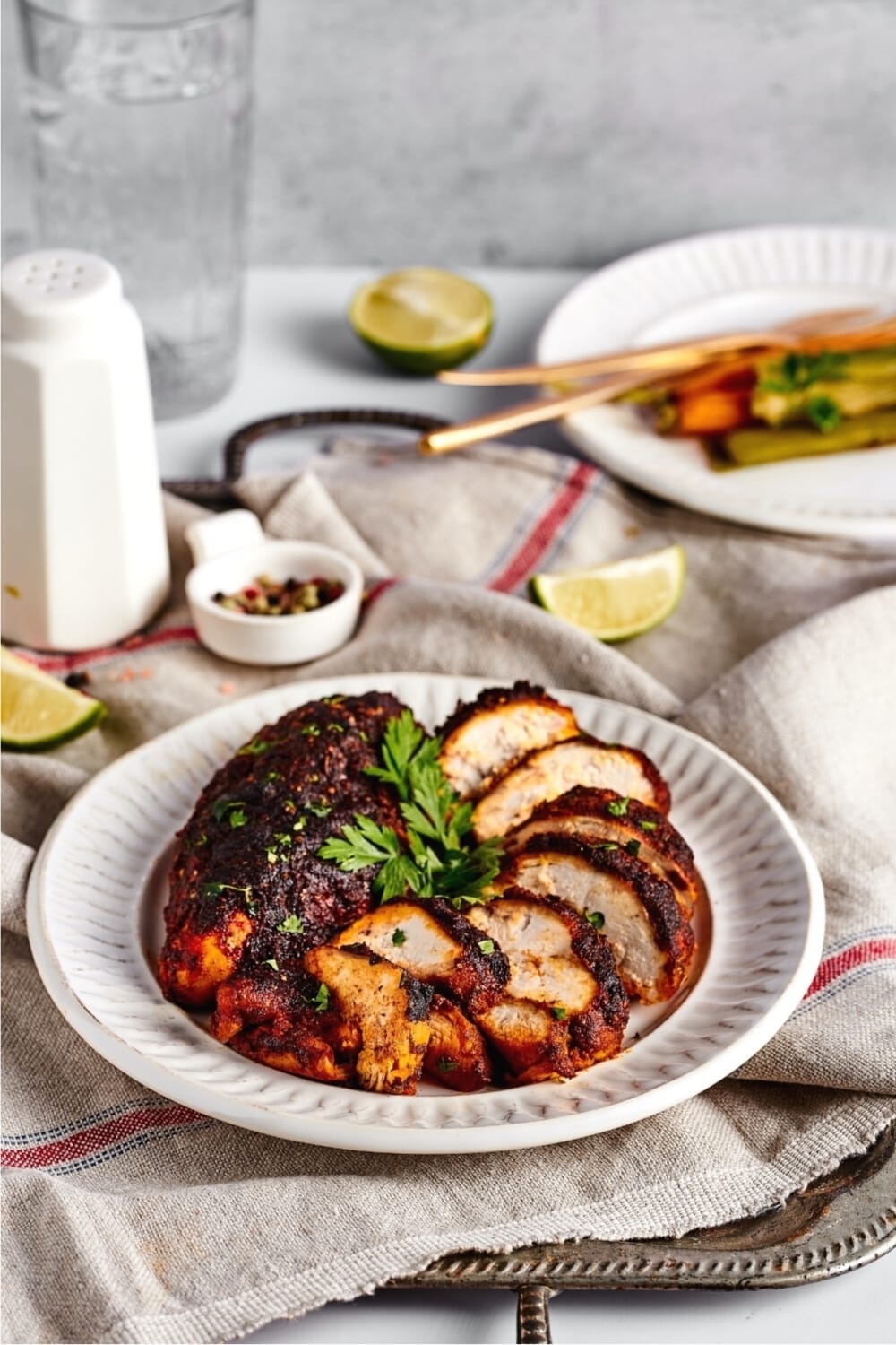 A white play with a whole piece of blackened chicken and a piece of blackened chicken cut into slices on it. Behind the plate as part of a white plate with carrots and leeks on it.