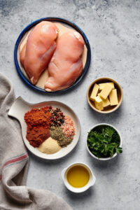 Small plate with two chicken breasts on a small bowl of cubed butter a tray of blackened chicken seasoning a small bowl of parsley and a small bowl of olive oil all on a gray counter.