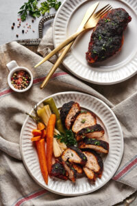 A white plate with a cut up piece of blackened chicken on it and some carrots and leeks next to it. Behind that is a white plate with a whole piece of blackened chicken and fork and knife on it. Both plates are on a gray tablecloth and a gray counter.