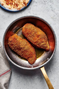 A skillet on a grey counter with two blackened chicken breasts in it.