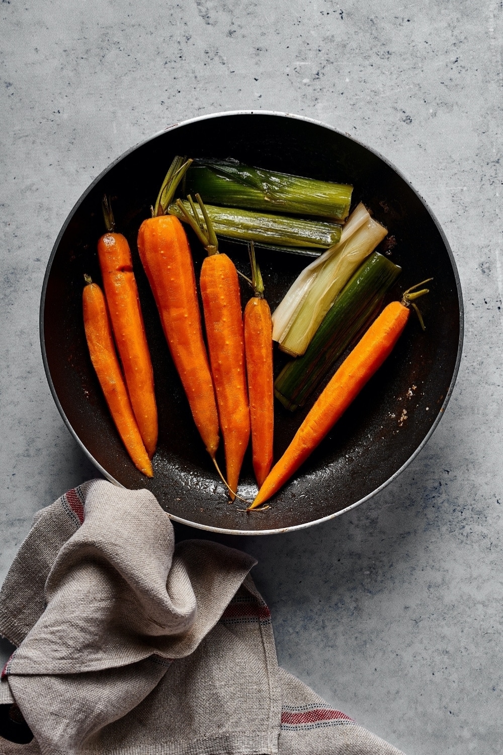 A skillet on a gray counter with carrots and leaks in it.