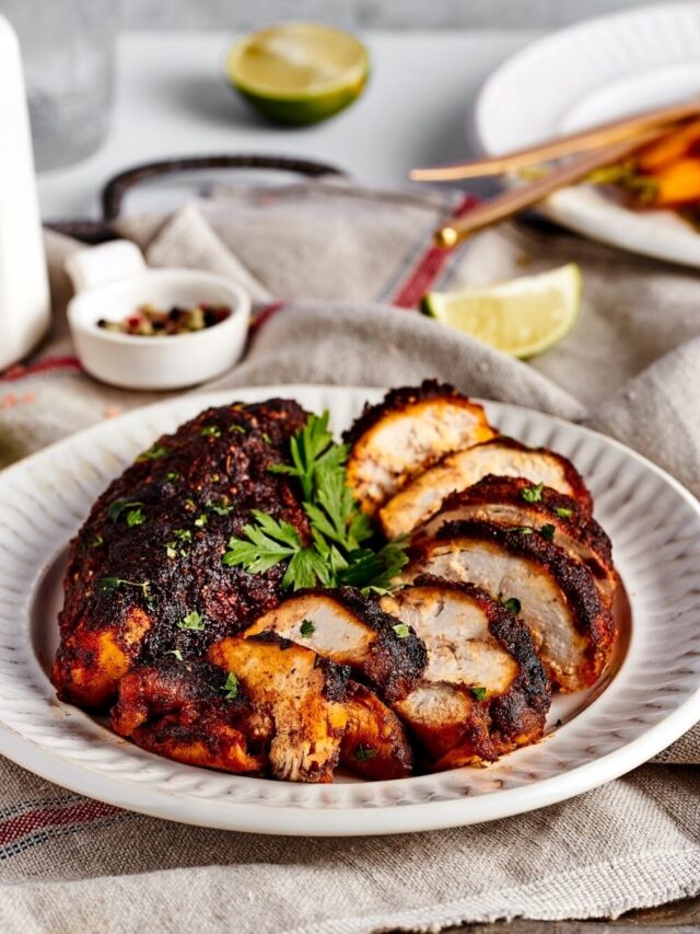 A white plate with a whole piece of blackened chicken breast and a sliced piece of blackened chicken on it.