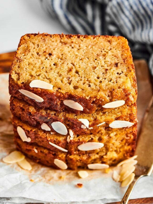 Four slices of banana bread stacked on top of one another on a piece of parchment paper. There are some sliced almonds in front of the Stack and part of a serving knife next to it.