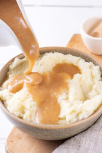 A gray bowl with mashed potatoes in it on a wooden cutting board. Part of a ladle is hovering over the bowl pouring gravy onto the mashed potatoes.