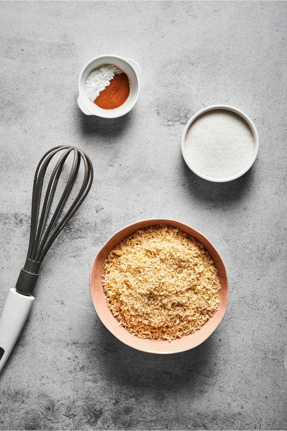 Small bowl of cinnamon and baking powder, a small bowl of confectioners erythritol, a small bowl of almond flour, and a whisk on a gray counter