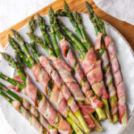A white plate with asparagus wrapped bacon on it. The plate is on a wooden cutting board on a white counter.
