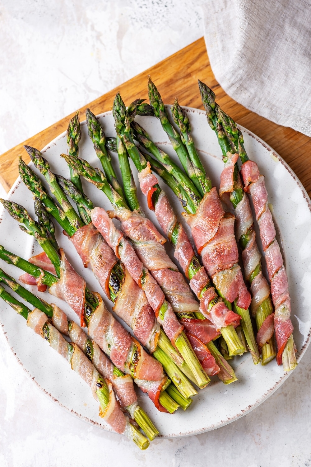 A white plate with asparagus wrapped bacon on it. The plate is on a wooden cutting board on a white counter.