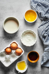 A small bowl of melted butter, a small bowl of milk, sour cream, banana extract, maple syrup, and three eggs all on a gray counter.