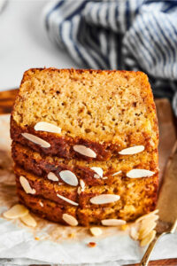 Four slices of banana bread stacked on top of one another on a piece of parchment paper. There are some sliced almonds in front of the Stack and part of a serving knife next to it.