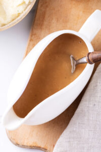 A ladle filled with gravy on a wooden cutting board. There is a spoon submerged in the gravy.