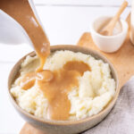 A gray bowl filled with mashed potatoes and a wooden cutting board. There is a ladle pouring gravy on to the mashed potatoes.