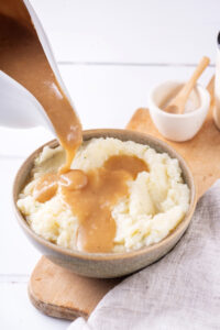 A gray bowl filled with mashed potatoes and a wooden cutting board. There is a ladle pouring gravy on to the mashed potatoes.