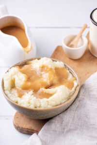 A gray bowl of mashed potatoes with gravy on top on a wooden cutting board. Behind the cutting board on a white counter is a ladle filled with gravy.