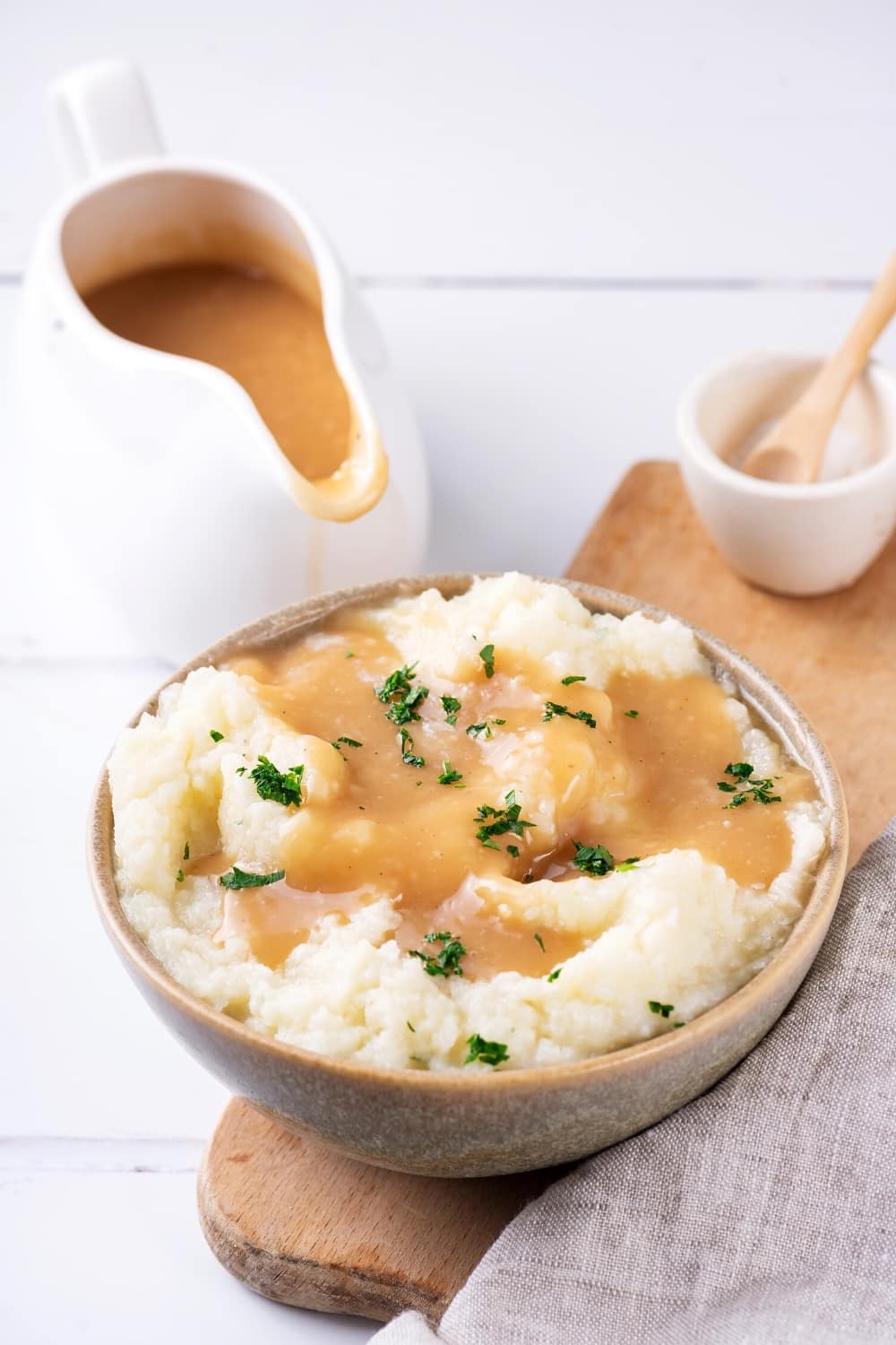 A bowl of mashed potatoes covered in gravy on a wooden cutting board. Behind the bowl on the white counter is a ladle filled with gravy.