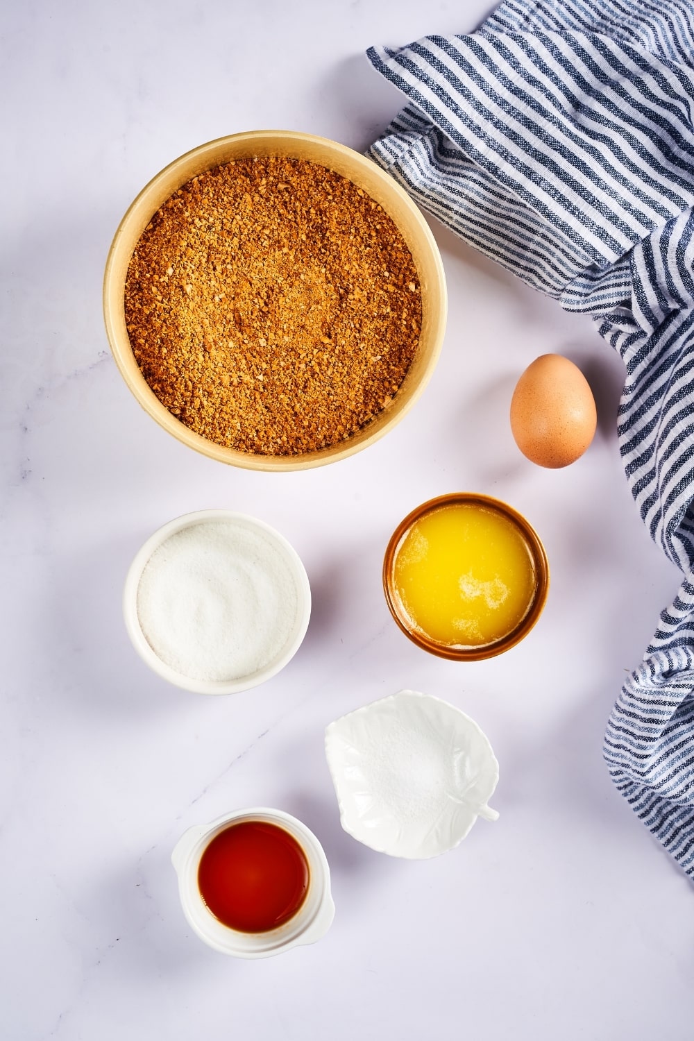 A large bowl filled with almond flour, one egg, a small bowl of melted butter, small bowl confectioners swerve, a small bowl salt, a small bowl of vanilla extract all on a white counter.