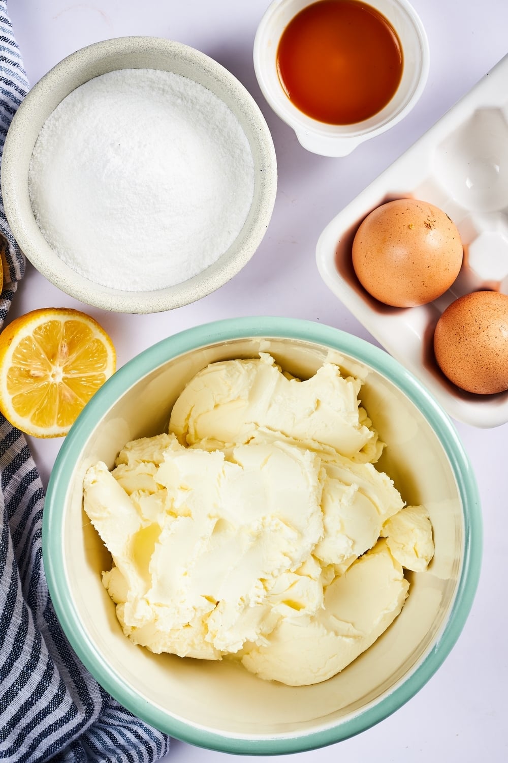 A large bowl filled with cream cheese. Behind it is a medium bowl filled with confectioners swerve, a small bowl of vanilla extract, two eggs, and a half a lemon. Everything is on a white counter.