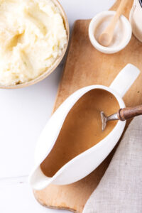 A ladle filled with gravy with a spoon in it. The ladle is on a wooden cutting board and next to it is part of a bowl with mashed potatoes in it.