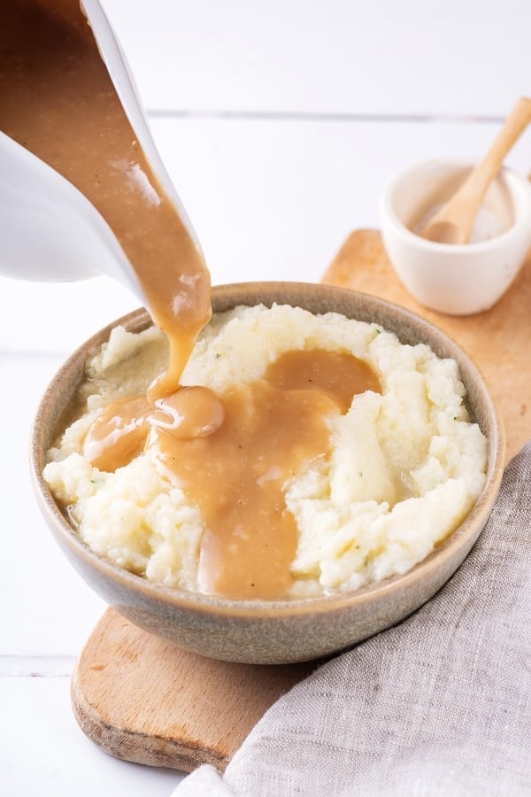 A gray bowl filled with mashed potatoes and a wooden cutting board. A ladle is pouring gravy onto the mashed potatoes.