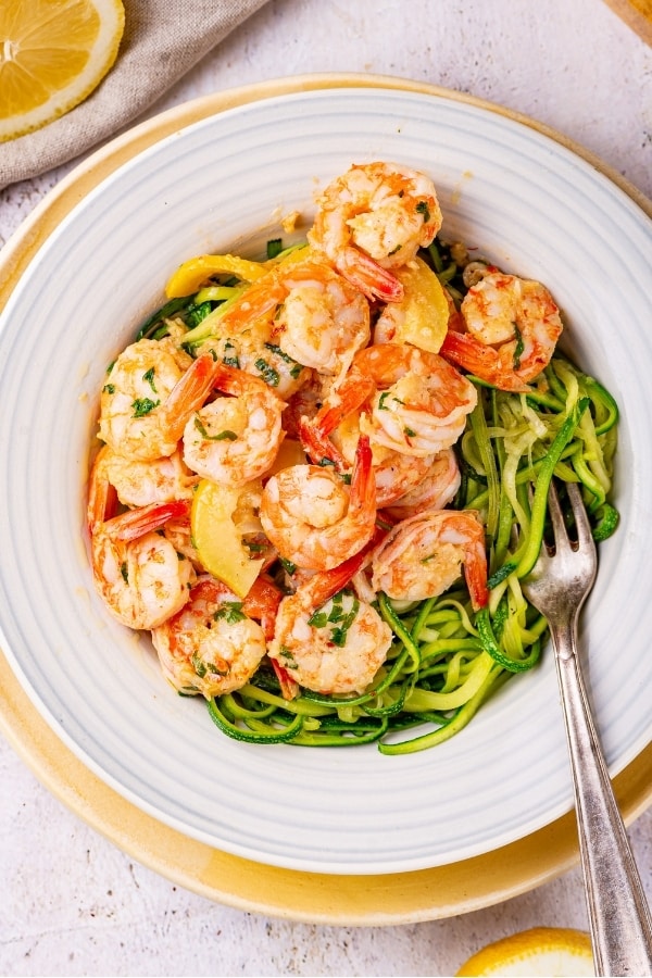 A white bowl filled with shrimp scampi on zoodles. The bowl is on a white plate on a white counter.