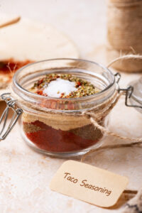 A glass jar with taco seasoning in it. There is a brown label that says "taco seasoning" on it in front of the jar.