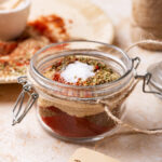 A glass jar with taco seasoning in it. In front of the jar is a brown label that says "taco seasoning" on it. And behind the jar is part of a white plate with taco seasoning mixture on it.