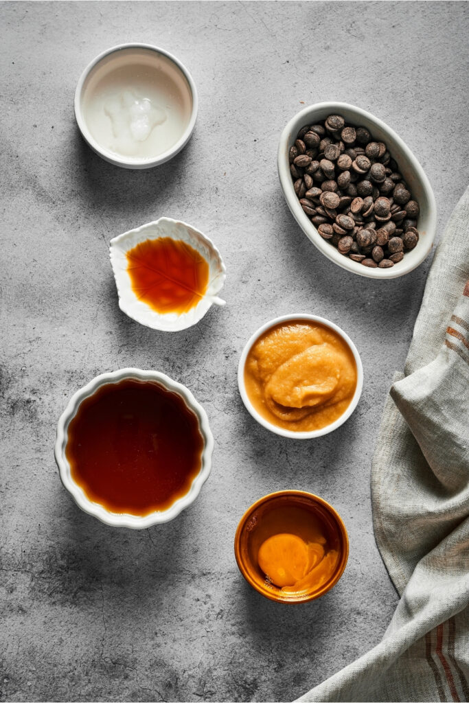 A small white bowl of baking powder, an oval bowl of chocolate chips, a small white leaf bowl with vanilla extract, a small white bowl filled with applesauce, a small bowl filled with an egg, and a medium bowl filled with honey. Everything is on a gray counter.