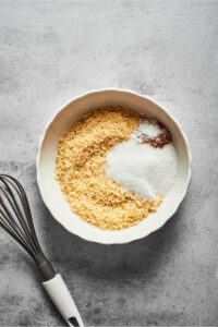 A small white bowl with almond flour, baking powder, cinnamon, and erythritol on a gray counter. There is a whisk to the left of the bowl.