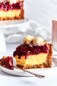 A slice of cheesecake on a white plate. There is a fork in front of the cheesecake with part of the cheesecake on it. Behind it is part of the cheesecake on a white serving tray.