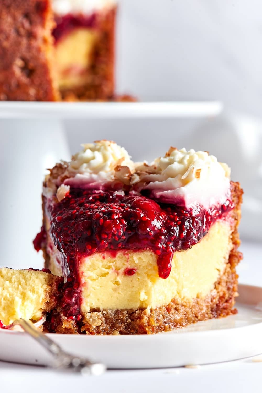 A slice of cheesecake on a white plate that is on my counter. A fork is touching the left the cheesecake and there is a piece from the front of the slice of the cheesecake on the prongs of the fork.