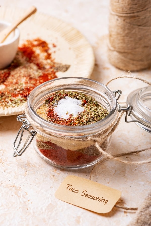 A glass jar filled with taco seasoning ingredients. Behind the jar is a white plate with the taco seasoning mixture on it and in front of the jar is a brown label that says "taco seasoning."