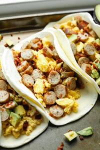 A baking sheet filled with breakfast tacos. The tacos have eggs, sausage, and bacon in them. Some of them have avocado and Sriracha as well.