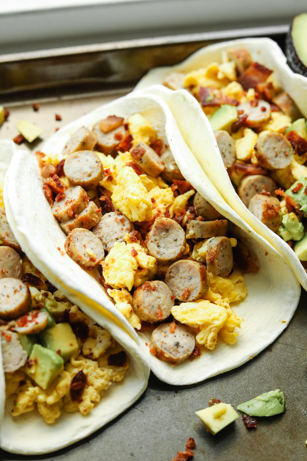 A baking sheet filled with breakfast tacos. The tacos have eggs, sausage, and bacon in them. Some of them have avocado as well.