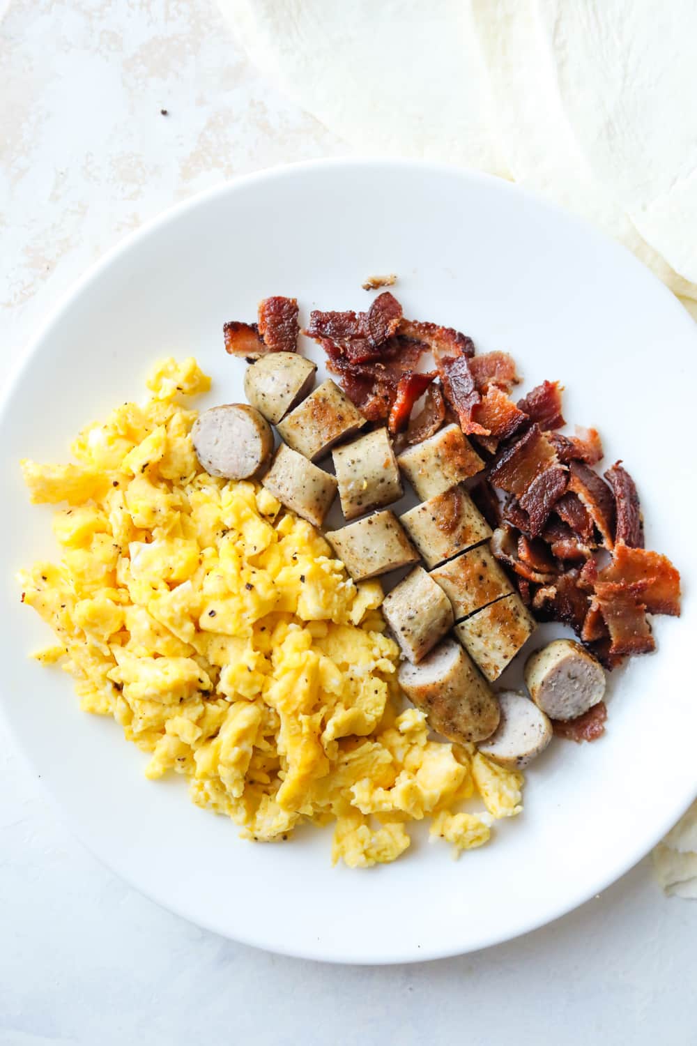 A white plate that is full of scrambled eggs and chopped bacon & sausage.